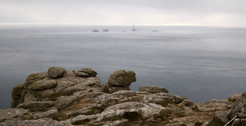 Misty longships lighthouse rocks This is a landscape photograph taken in the early evening during the summer season, featuring the misty Longships lighthouse rocks as the main subject. The image captures the rugged cliffs of Cornwall in the foreground, overlooking the expansive and calm sea that stretches out towards the horizon. The Longships Lighthouse, located on the distant rocks, is visible through a veil of mist, emphasizing the atmospheric conditions typical of Cornwall’s coastline. The rocky terrain of the cliffs and the serene sea together highlight the characteristic natural beauty of this region.
